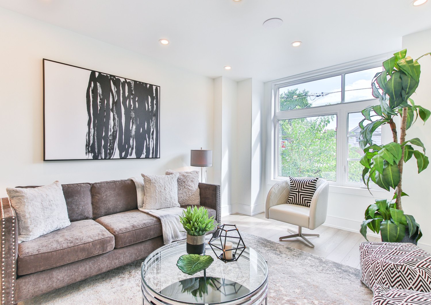 A furnished living room with a corner sofa, a low glass table and a tall window looking onto greenery